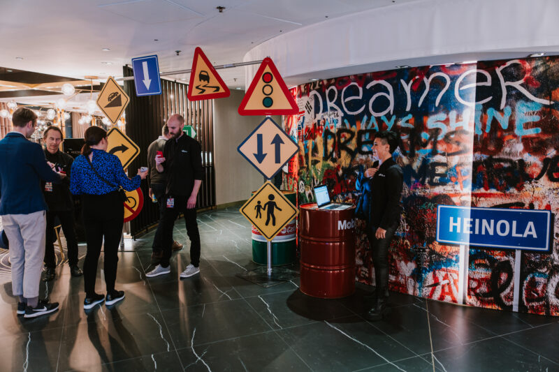 Liikennemerkeistä tehdyn portin alla seisoo mies, joka keskustelee naisen kanssa. Vieressä on graffitiseinä, jonka edessä öljytynnyrin vieressä seisoo nainen. Öljytynnyrin päällä on kannettava tietokone.