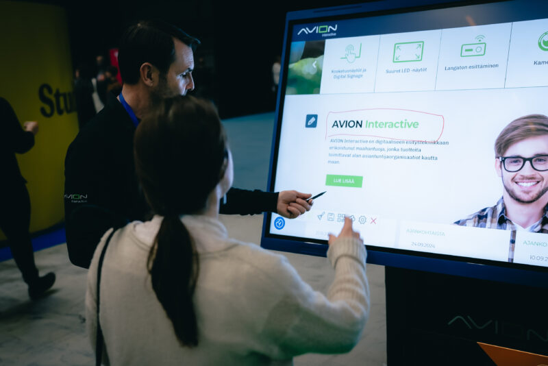 Mies esittelee naiselle näytöllä olevaa infoa. Näytöllä on ympyröitynä teksti AVION Interactive.