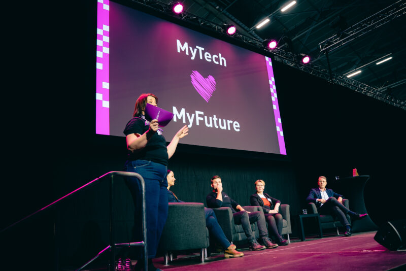 Lavalla seisoo nainen puhumassa. Hänen takanaan istuu neljä henkilöä rivissä nojatuoleissa. Seinällä on suuri näyttö, jossa lukee MyTech MyFuture.